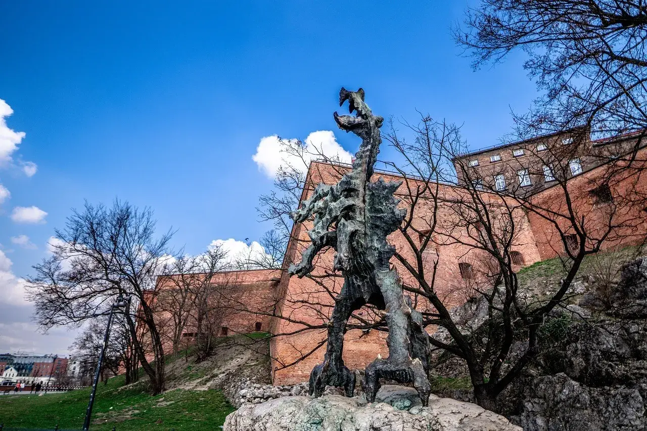 Smok Wawelski Kraków, groźny posąg smoka u stóp Wawelu, na tle ceglanych murów i błękitnego nieba.