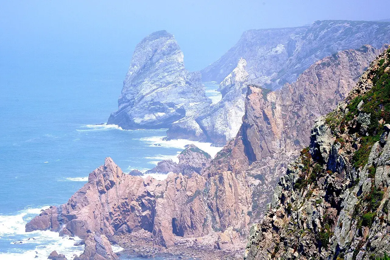 Skaliste wybrzeże Cabo da Roca, z falami rozbijającymi się o klify, w oddali widać mgliste góry.