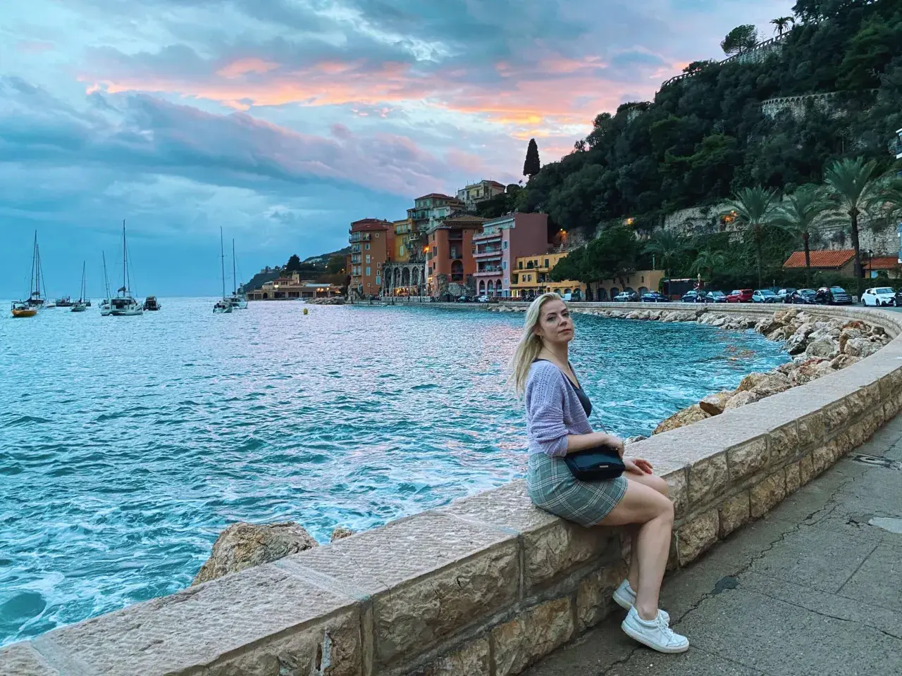 Kobieta siedzi na kamiennym murku nad morzem w Villefranche-sur-Mer, podziwiając zachód słońca i żaglówki.