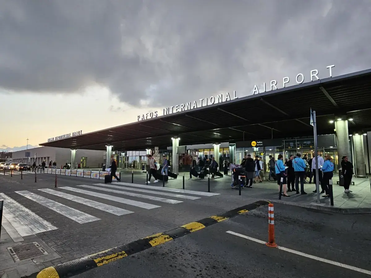 Ludzie z bagażami wchodzą do portu lotniczego Pafos pod neonowym napisem.