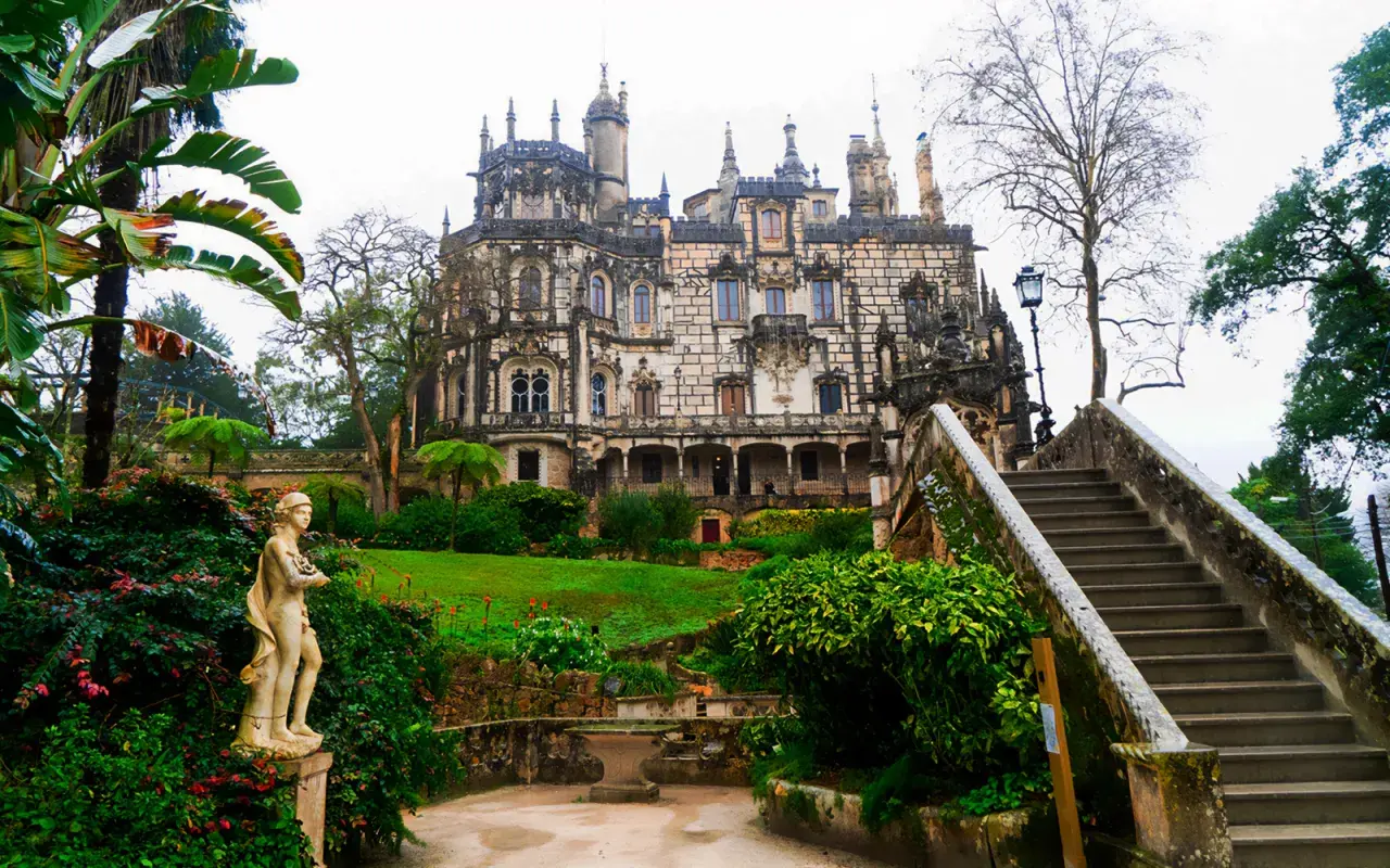 Kamienna rzeźba w ogrodzie Quinta da Regaleira, z widokiem na ozdobny pałac i schody prowadzące do niego.