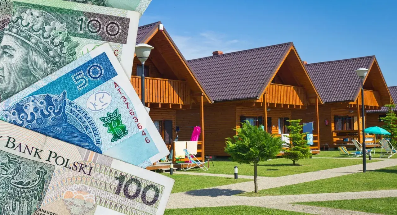 Polskie banknoty przed drewnianymi domkami letniskowymi. Zastanawiasz się, gdzie najlepiej rezerwować noclegi, by wakacje były udane i nie zrujnowały budżetu?