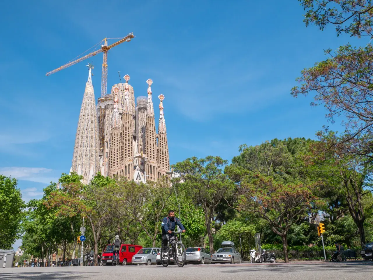 Majestatyczna La Sagrada Familia w Barcelonie, wciąż w budowie, góruje nad zielonym parkiem i ulicą, gdzie rowerzysta przemierza drogę.