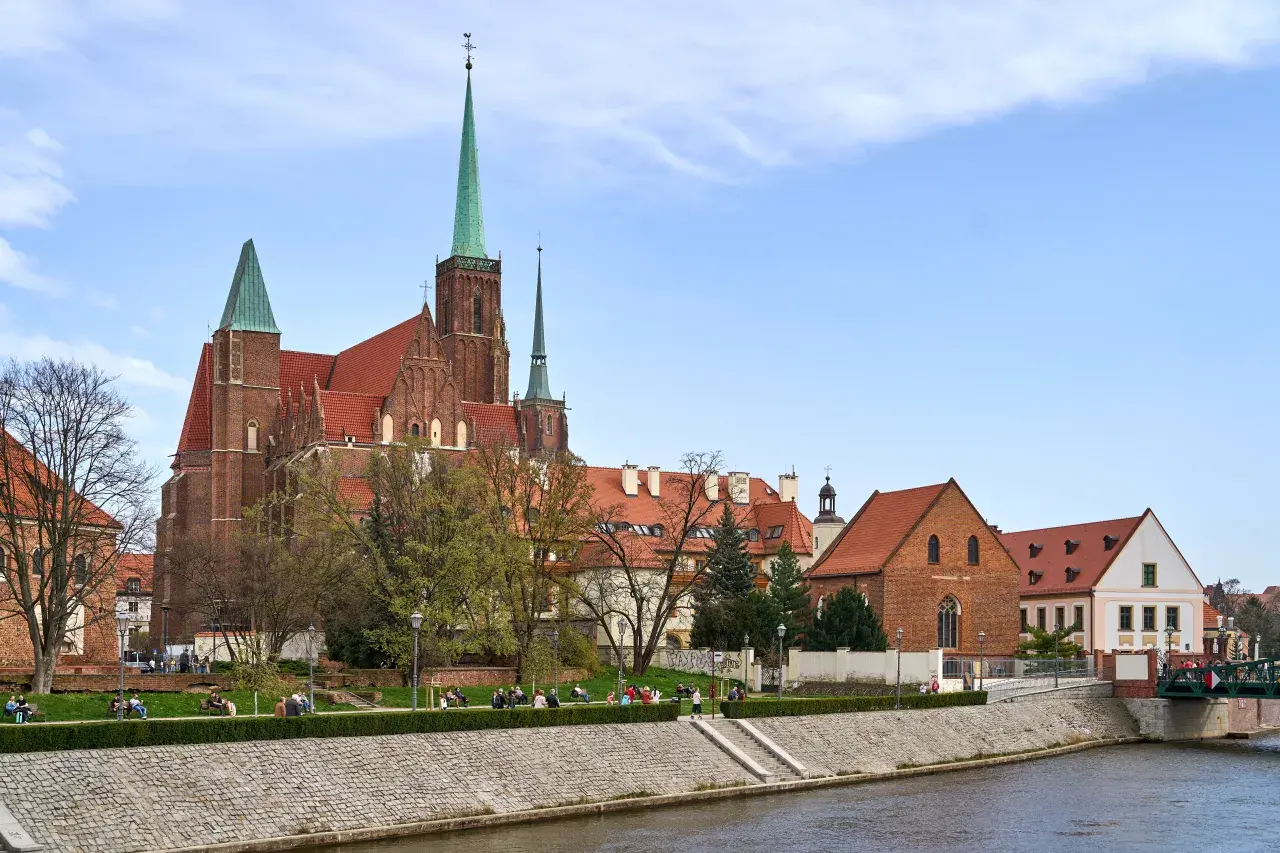Ostrów Tumski we Wrocławiu z katedrą i zielonymi wieżami, nad rzeką, z ludźmi spacerującymi po bulwarze.