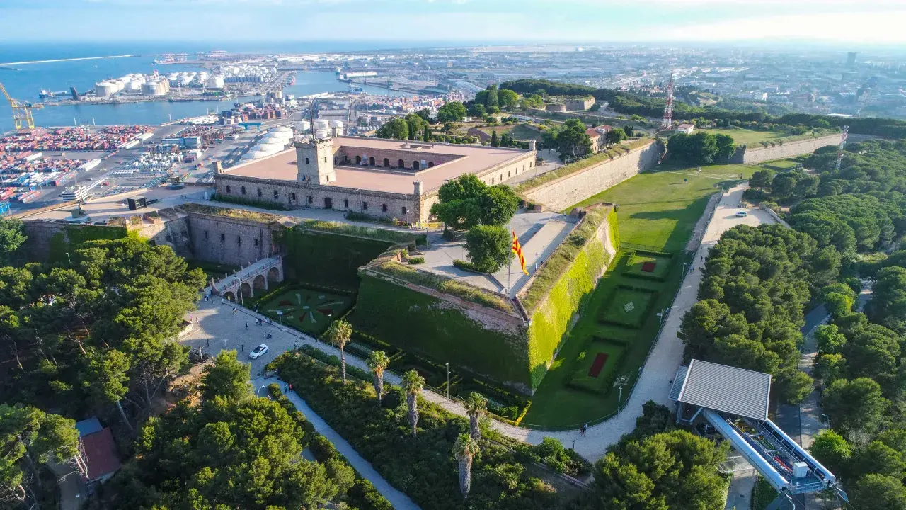 Zamek Castell de Montjuïc góruje nad portem w Barcelonie. Otoczony zielenią, z flagą Katalonii powiewającą na wietrze.