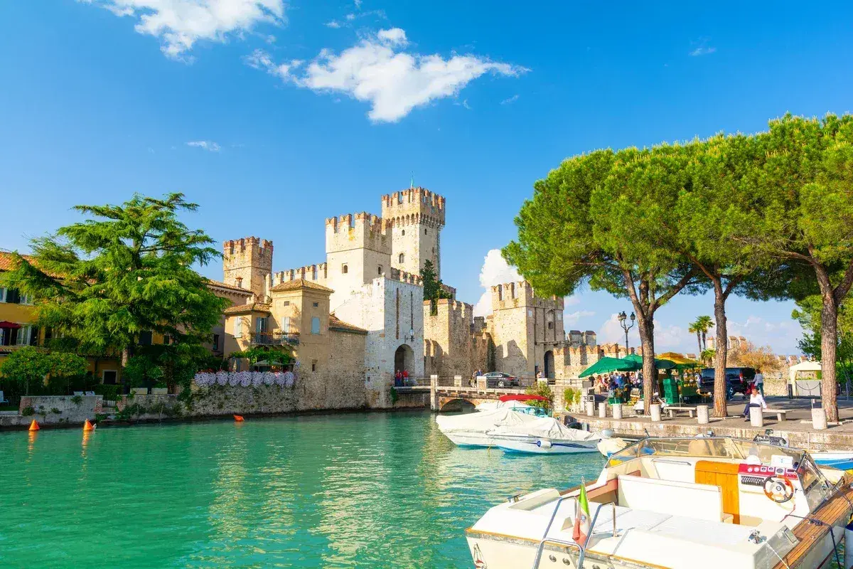 Malowniczy widok na Sirmione Castello Scaligero nad turkusową wodą, z łodziami zacumowanymi przy brzegu i zielonymi drzewami.
