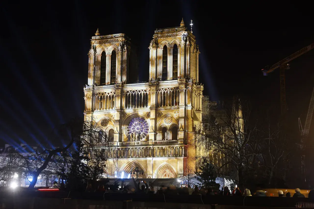 Katedra Notre Dame w Paryżu nocą, oświetlona reflektorami. Widać tłum ludzi oglądających spektakl.