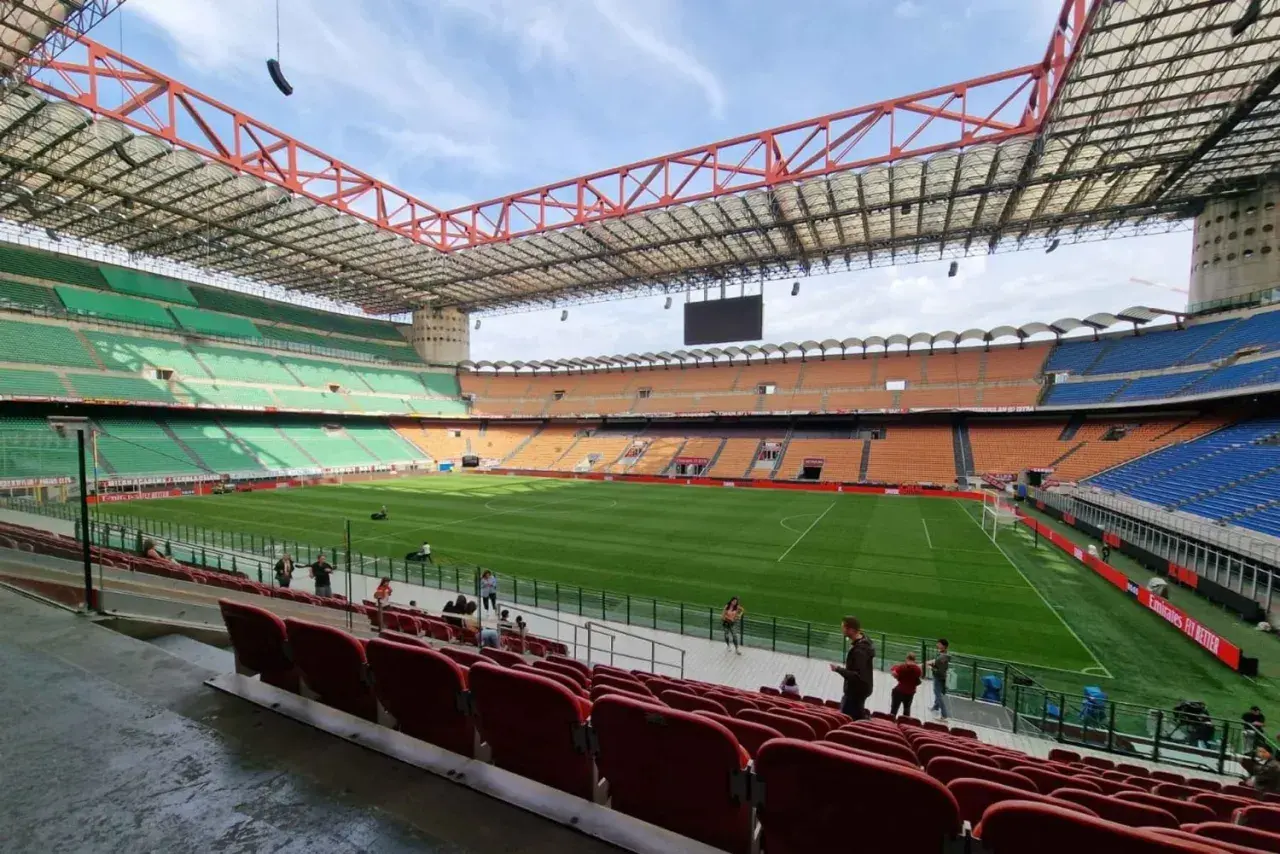 Pusta trybuna stadionu San Siro z zieloną murawą i pomarańczowymi oraz niebieskimi siedzeniami.