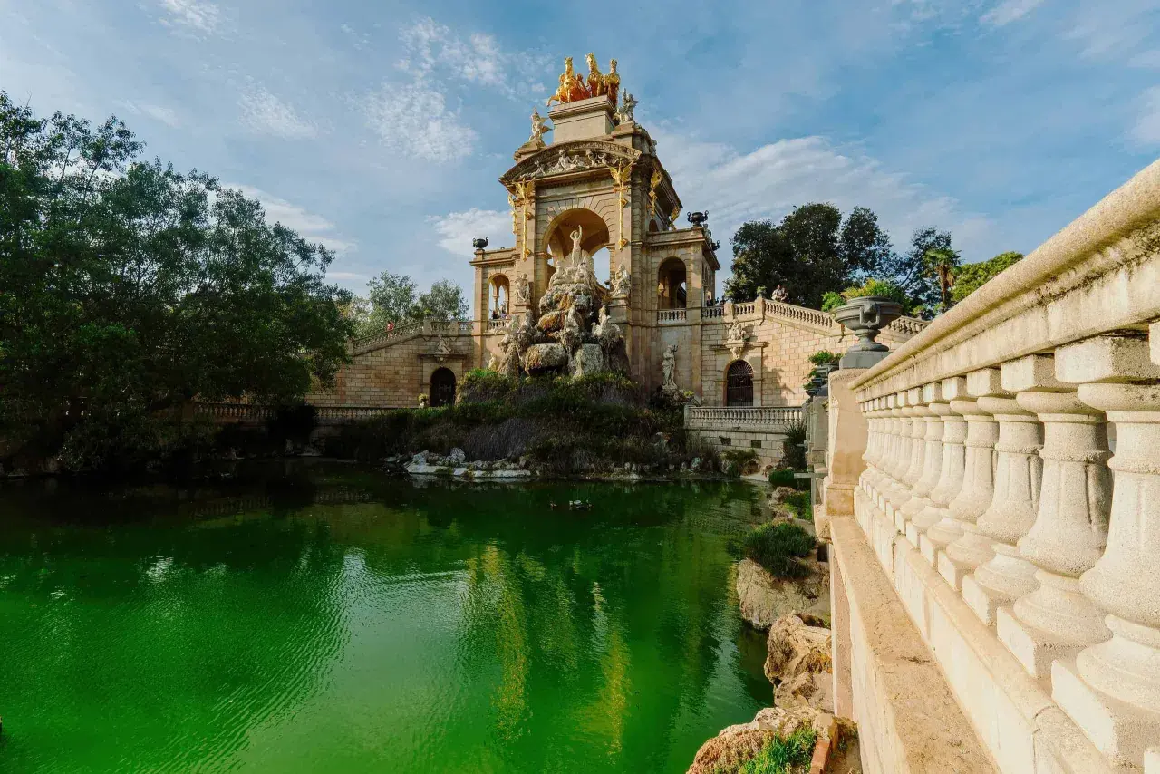 Monumentalna fontanna z rydwanem na szczycie, otoczona zielonym stawem i kamiennymi schodami w Parc de la Ciutadella.