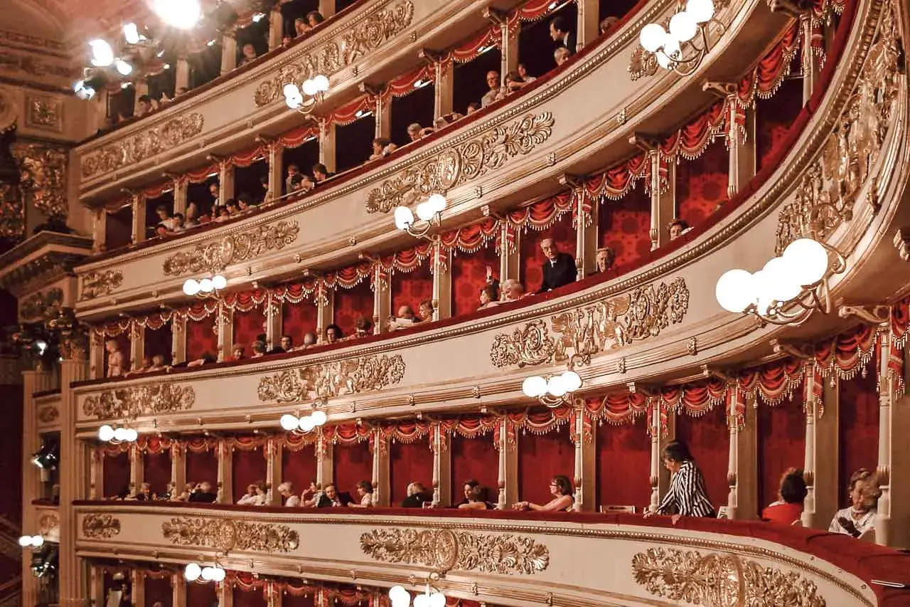 Wnętrze La Scala w Mediolanie, z widownią w lożach, ozdobione złotymi zdobieniami i czerwonymi zasłonami.