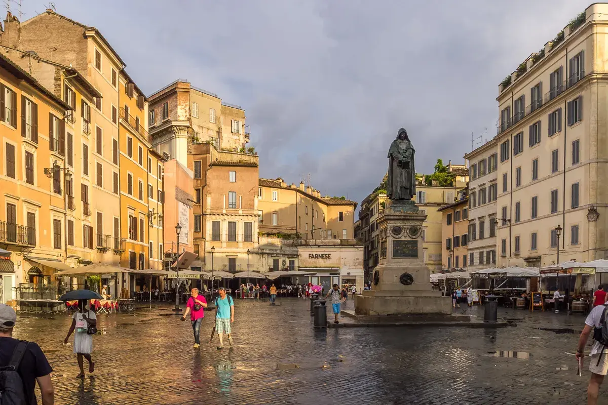 Pomnik na placu Campo de' Fiori w Rzymie, otoczony kolorowymi budynkami i ludźmi spacerującymi po mokrym bruku.