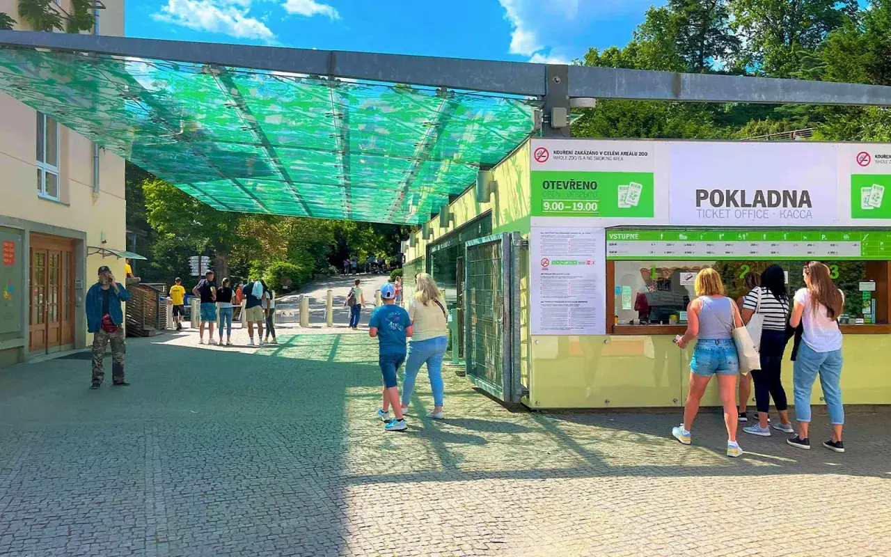 Wejście do ogrodu zoologicznego w Pradze. Ludzie stoją w kolejce do kasy biletowej pod zielonym zadaszeniem.