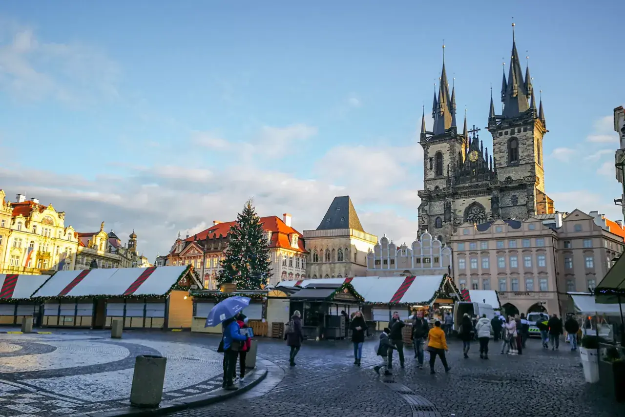 Rynek Staromiejski w Pradze, z choinką i straganami świątecznymi, podziwiany przez spacerujących ludzi.