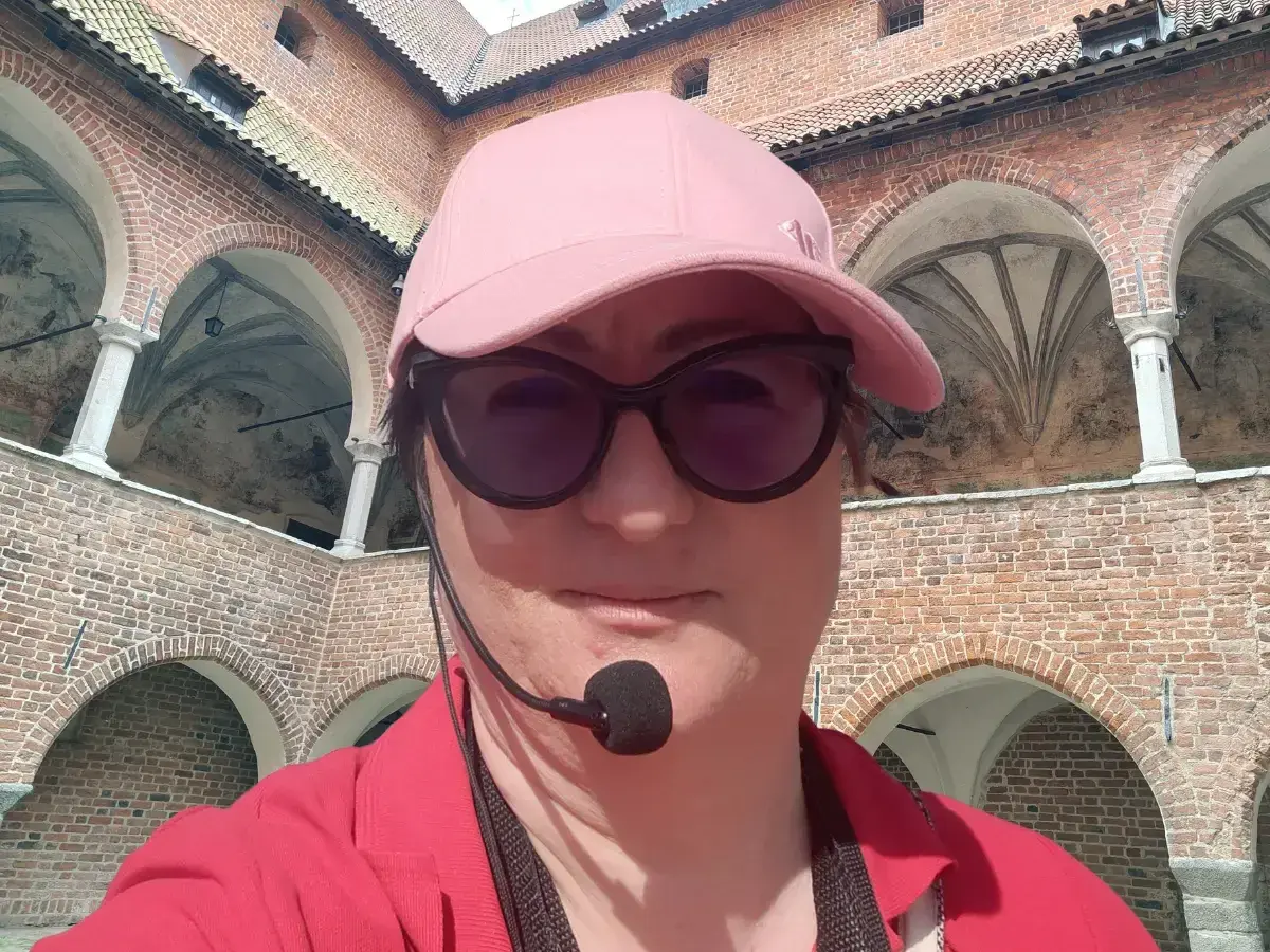 Kobieta w czapce i okularach przeciwsłonecznych zwiedza z przewodnikiem z mikrofonem zamek w Lidzbarku Warmińskim, odkrywając jego atrakcje.
