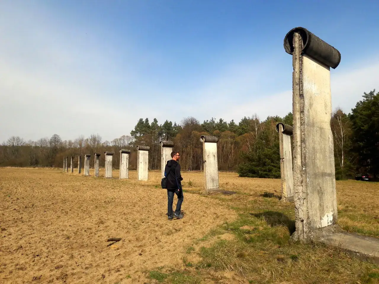 Fragment muru berlińskiego w krajobrazie. Człowiek spaceruje obok betonowych słupów, które kiedyś dzieliły miasto.