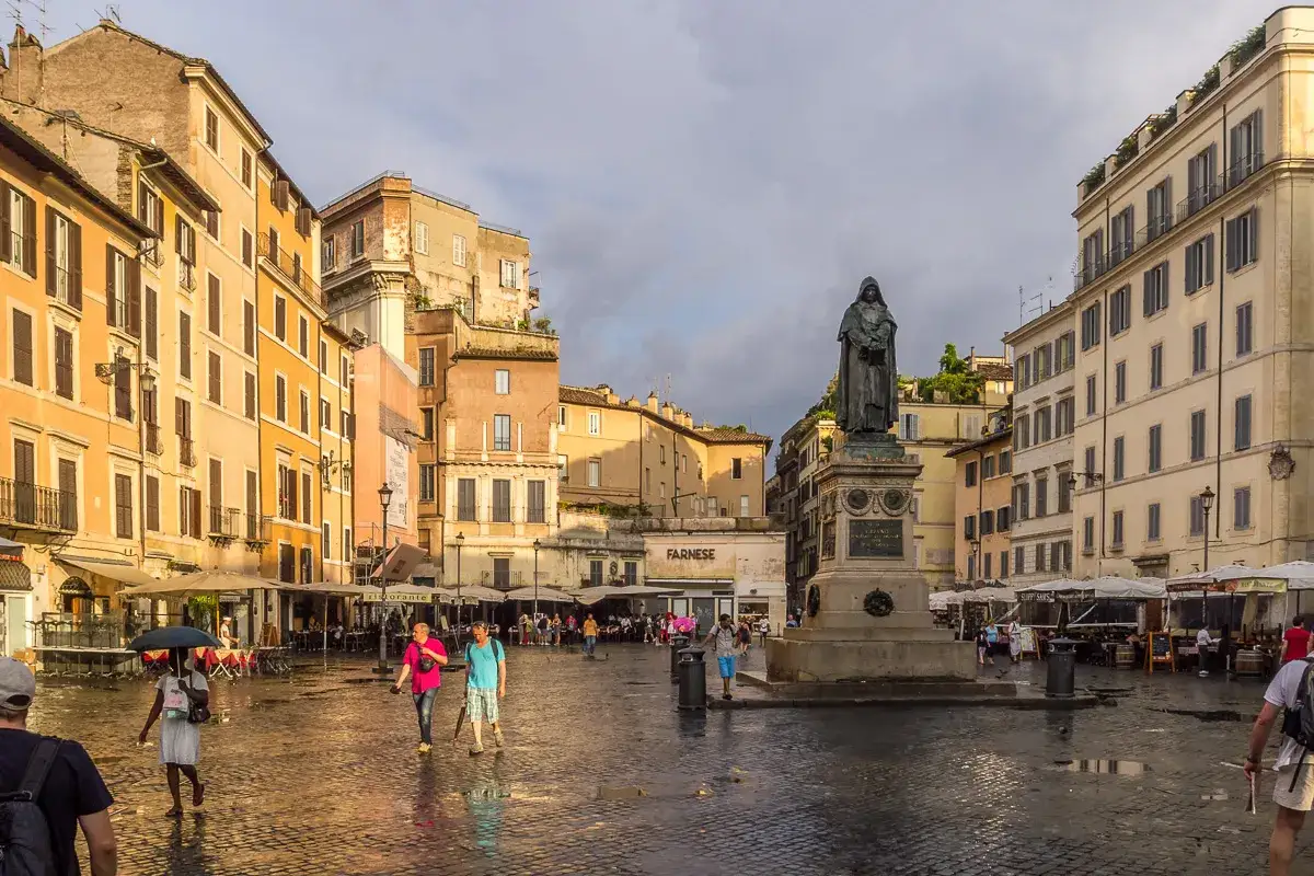 Pomnik na placu Campo de' Fiori w Rzymie, otoczony kolorowymi kamienicami i ludźmi spacerującymi po mokrym bruku.