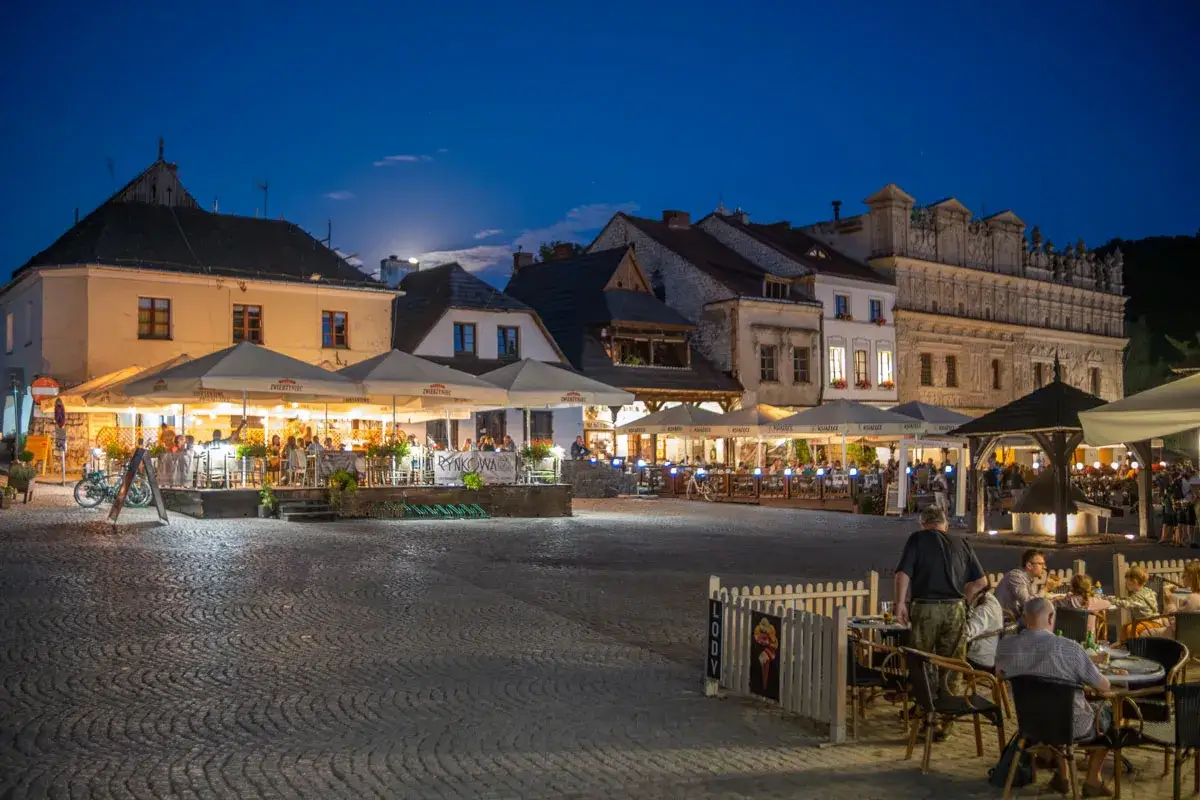 Wieczorny Rynek w Kazimierzu Dolnym tętni życiem. Stoliki restauracyjne pod parasolami, zabytkowa zabudowa i księżyc tworzą niepowtarzalne kazimierz dolny atrakcje.