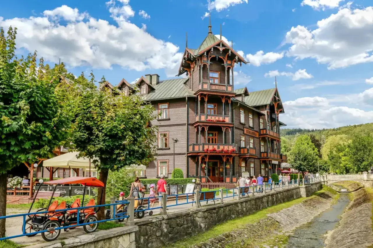 Drewniana willa w Krynicy Zdroju, z charakterystyczną architekturą, to jedna z atrakcji. Spacerowicze i rowerzyści cieszą się słonecznym dniem nad strumieniem.