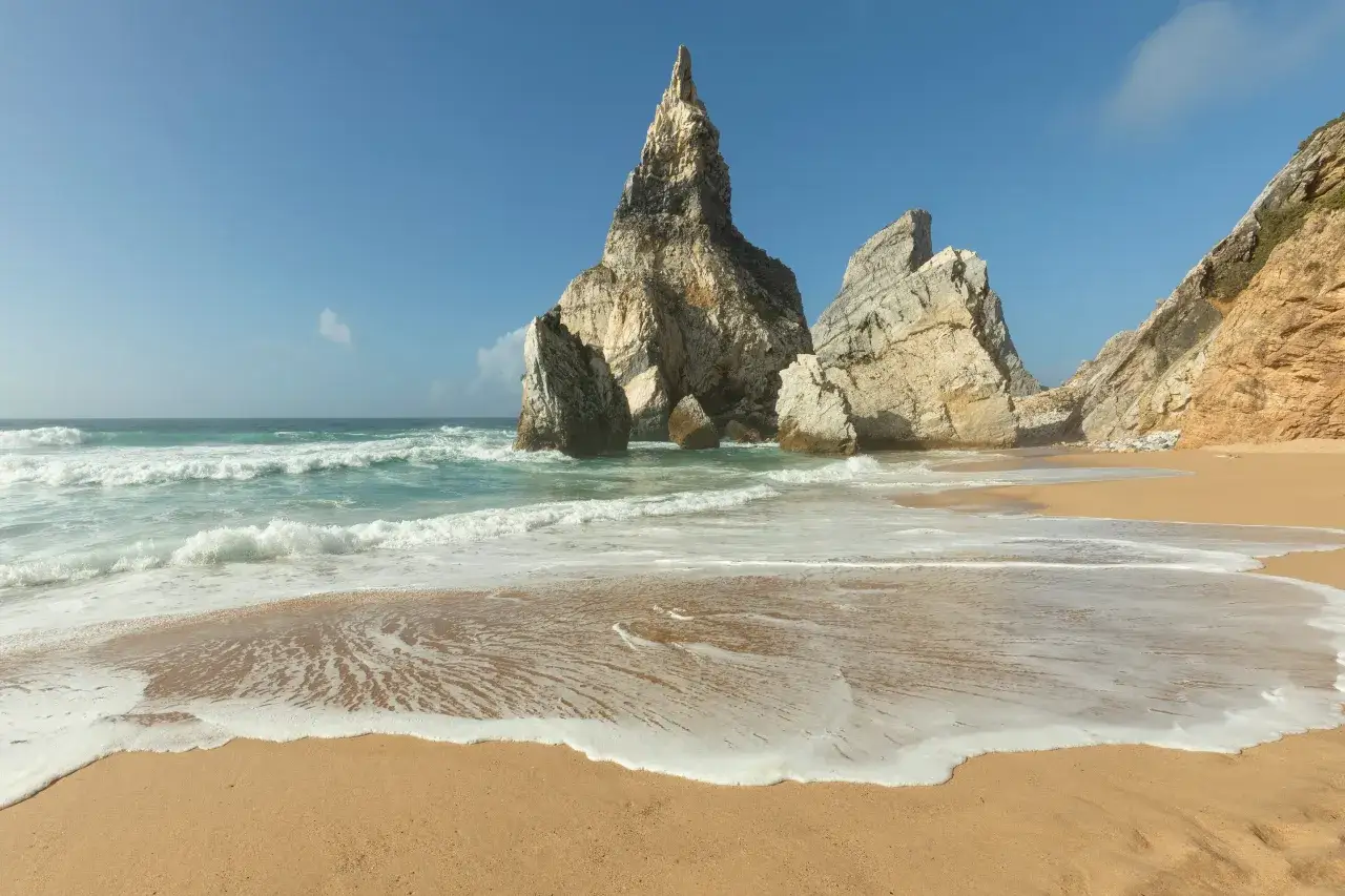 Plaża z piaskiem i skalnymi formacjami, gdzie fale oceanu obmywają brzeg. Widok na Cabo da Roca, najbardziej wysunięty na zachód punkt Europy.
