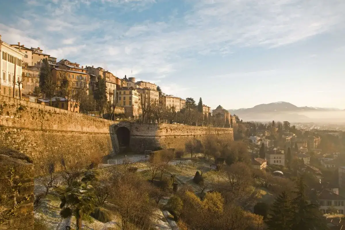 Widok na zabytkowe miasto Bergamo, z widocznymi murami obronnymi i budynkami. W oddali majaczą góry, a w tle widać mglisty krajobraz, który może sugerować bliskość lotniska Mediolan Bergamo.