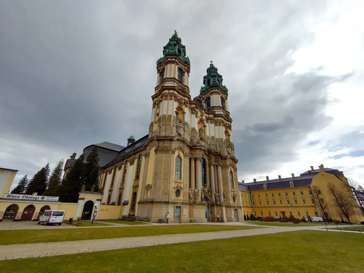 Majestatyczne opactwo pocysterskie w Krzeszowie z dwiema wieżami i żółtym budynkiem klasztornym na tle pochmurnego nieba.
