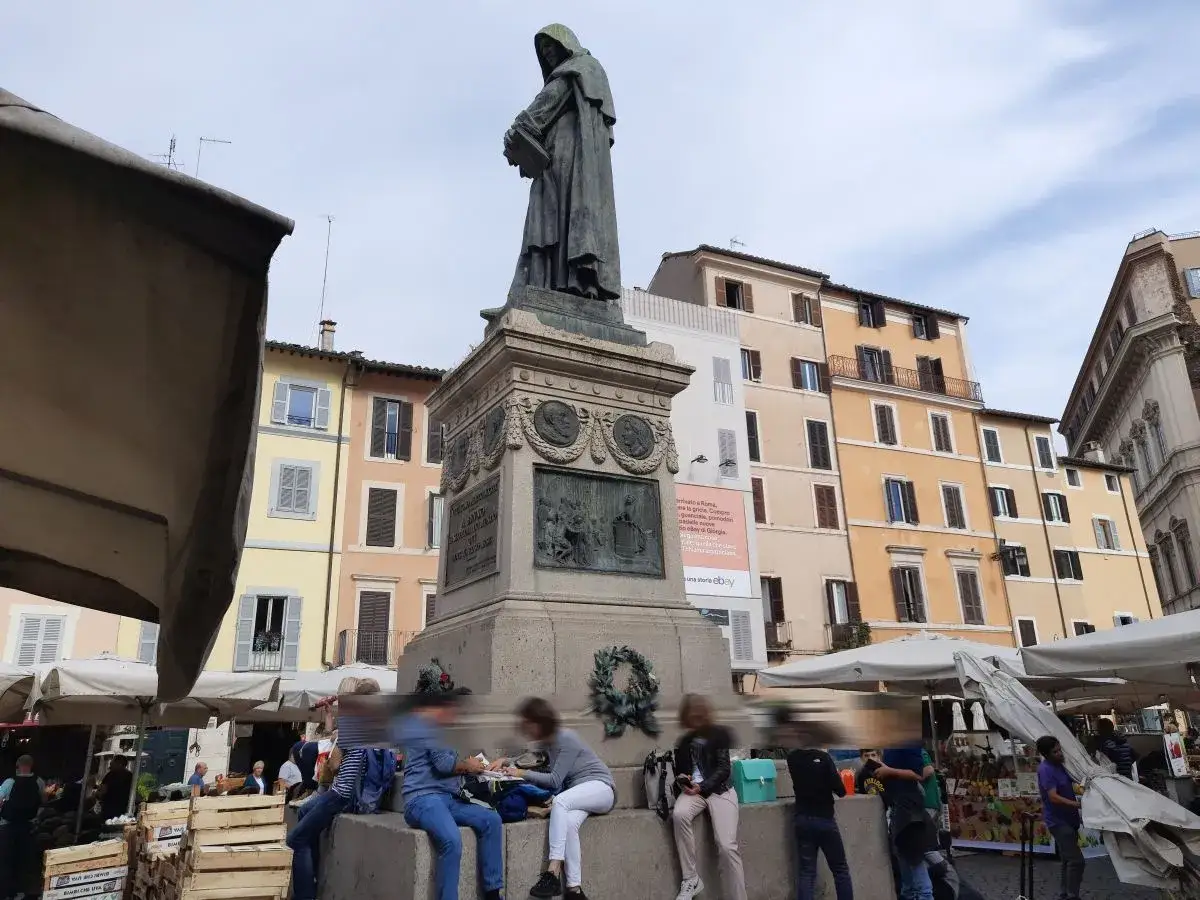 Pomnik na Campo de' Fiori w Rzymie, otoczony straganami i ludźmi.