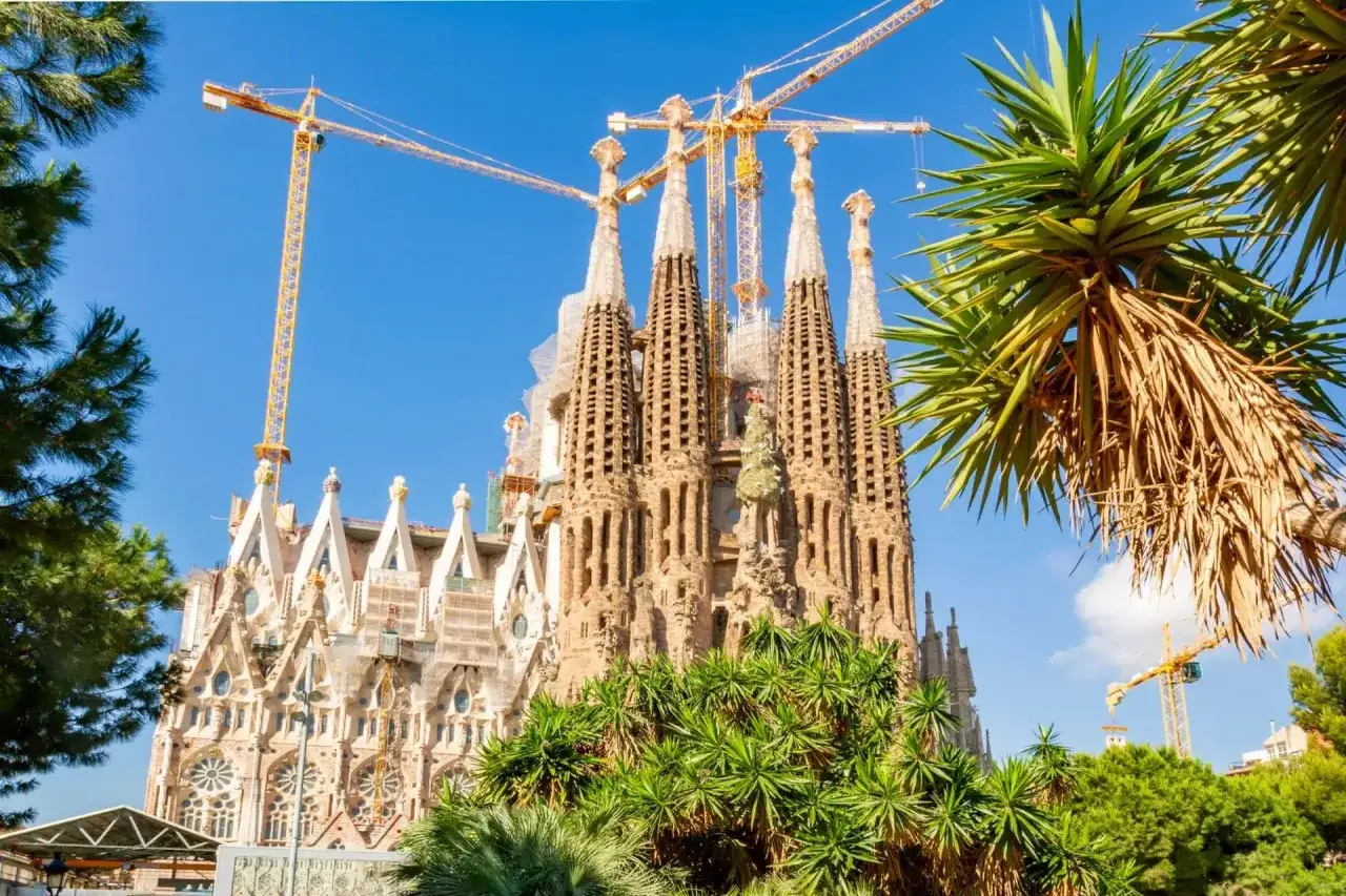 La Sagrada Familia w Barcelonie, wciąż w budowie, otoczona zielenią i górującymi dźwigami.