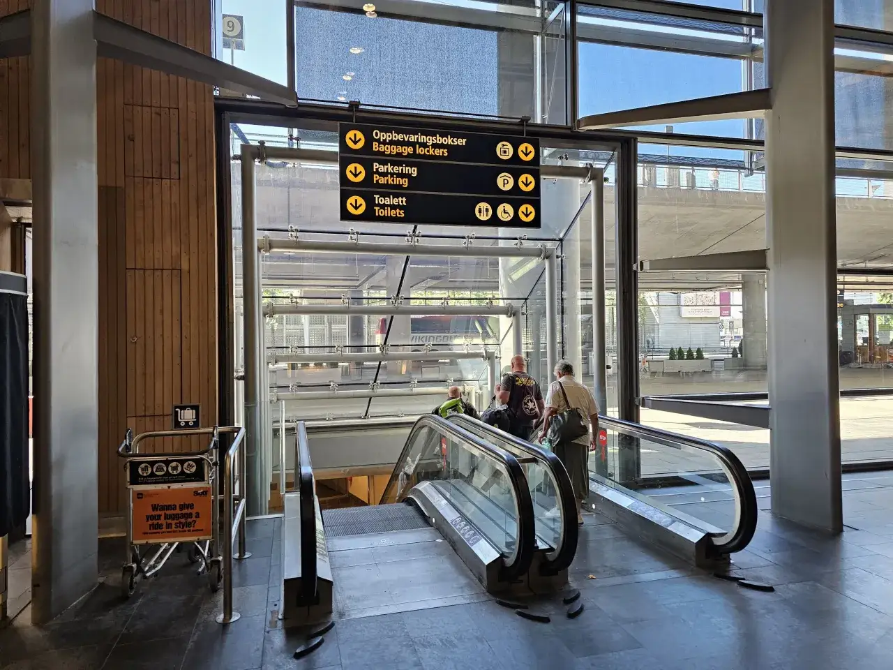 Schody ruchome na lotnisku Oslo Gardermoen. Znaki wskazują przechowalnię bagażu, parking i toalety.