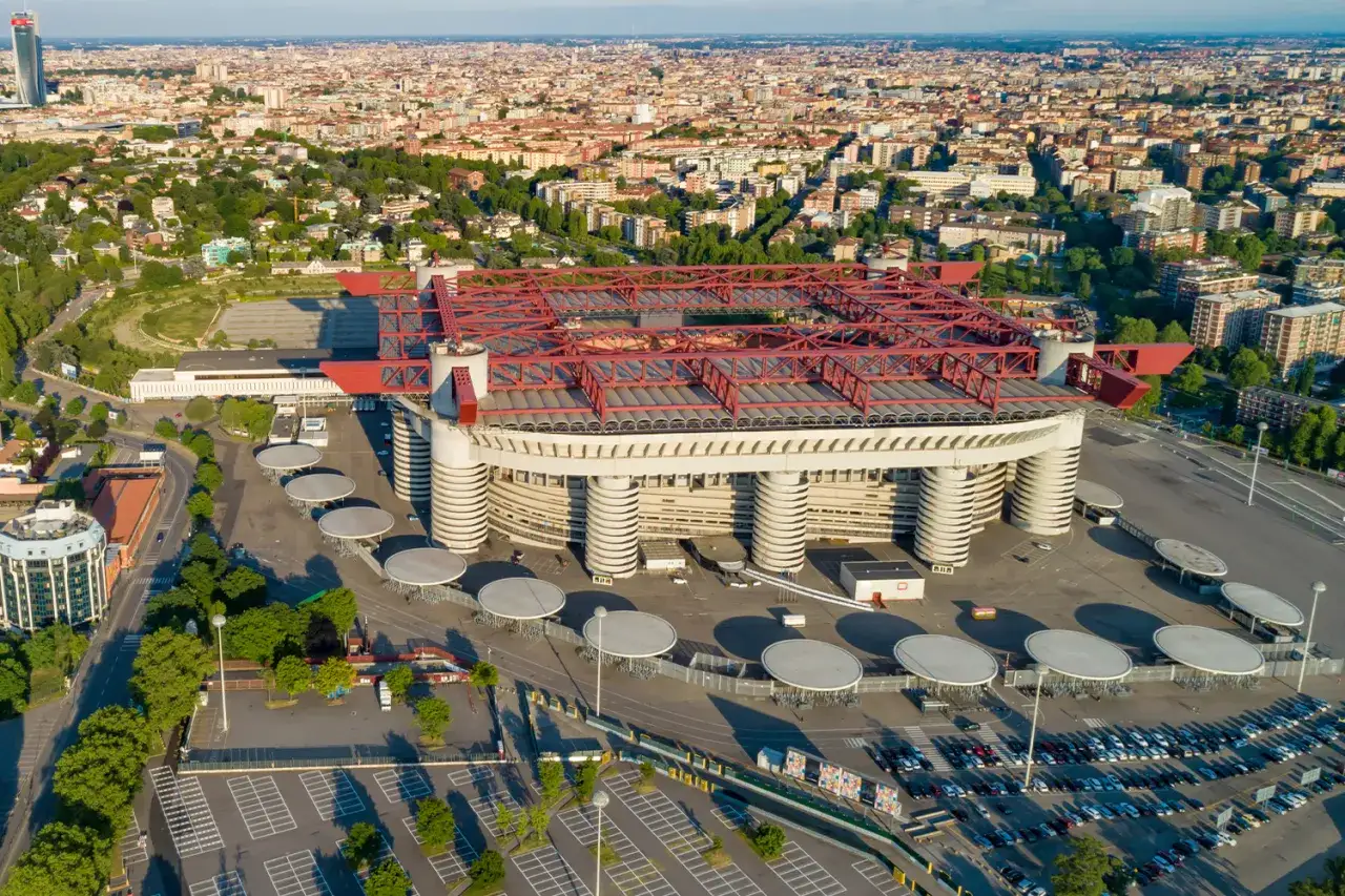 Stadion Interu Mediolan, znany jako San Siro, z charakterystyczną czerwoną kratownicą dachu i cylindrycznymi podporami, otoczony parkingami i zielenią miejską.