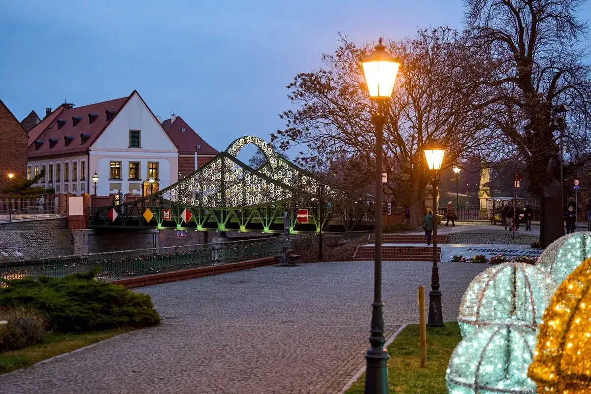 Oświetlony most na Ostrowie Tumskim we Wrocławiu, z zabytkowymi kamienicami i latarniami tworzy magiczny klimat.