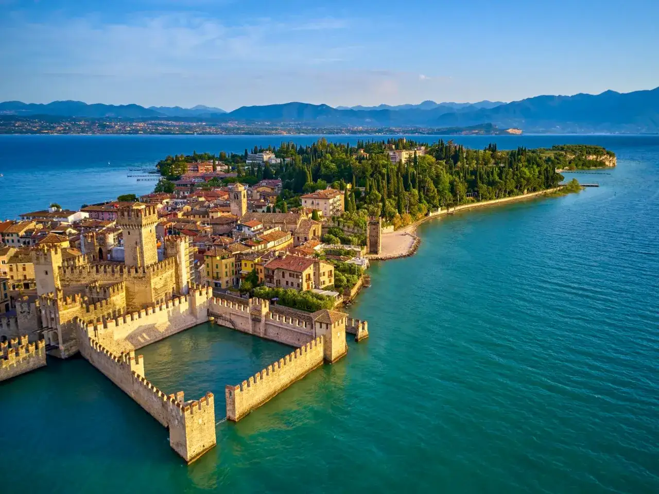 Malowniczy widok na Sirmione, z imponującym Castello Scaligero otoczonym wodą, na tle błękitnego jeziora i gór.
