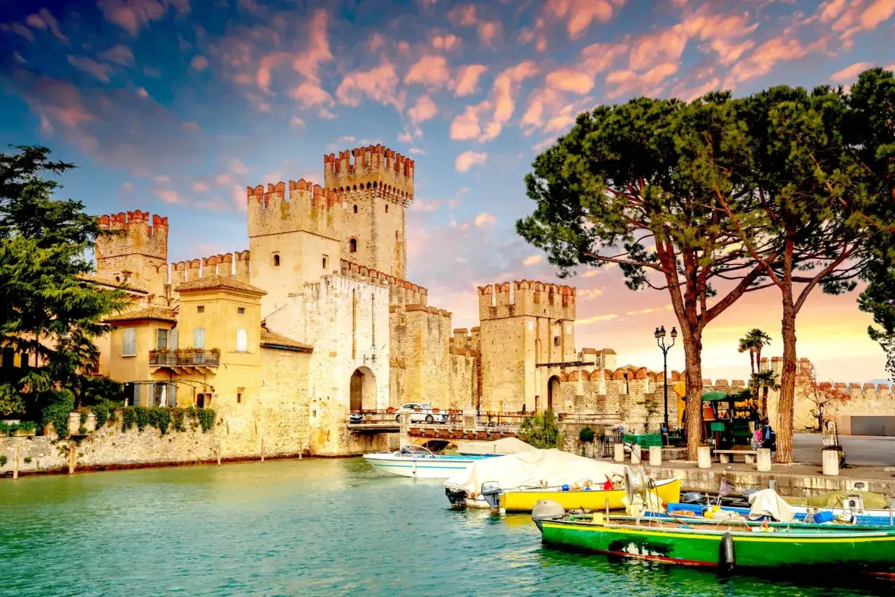 Zamek Scaligero w Sirmione nad jeziorem Garda, z łodziami przy brzegu i malowniczym niebem.