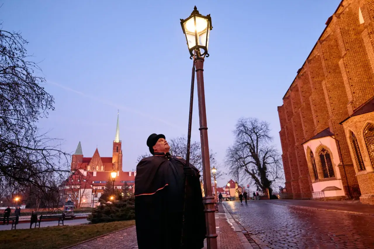 Latarnik na Ostrowie Tumskim we Wrocławiu zapala lampę. W tle katedra i zabytkowe kamienice.