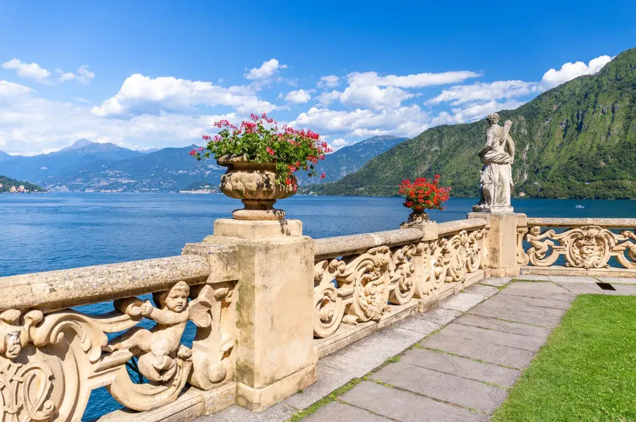 Kamienna balustrada z rzeźbami i kwiatami nad Lago di Como, w tle góry i błękitne niebo.