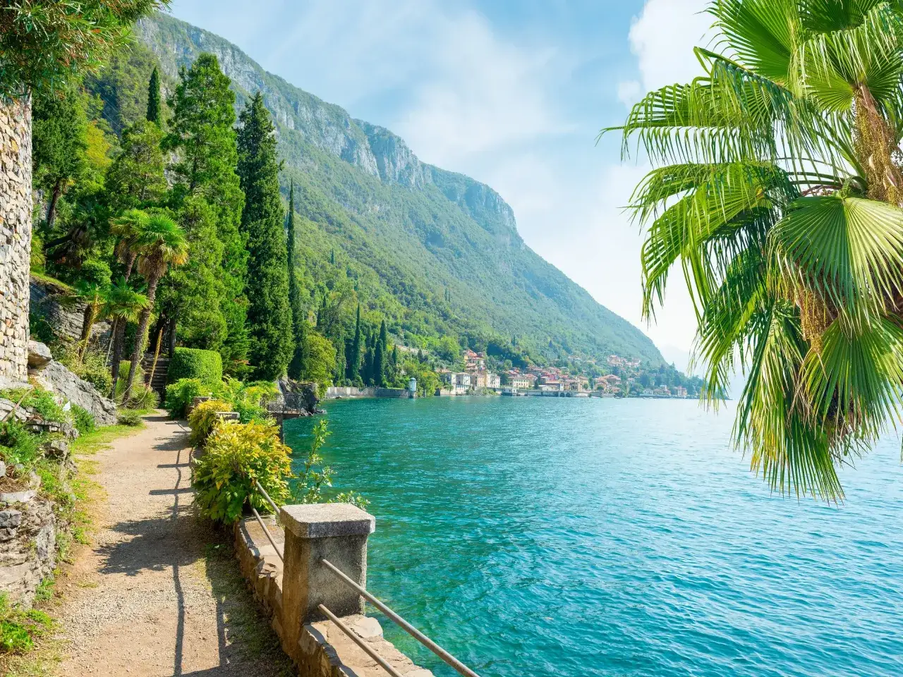 Malowniczy brzeg Lago di Como z palmami, cyprysami i górami w tle.