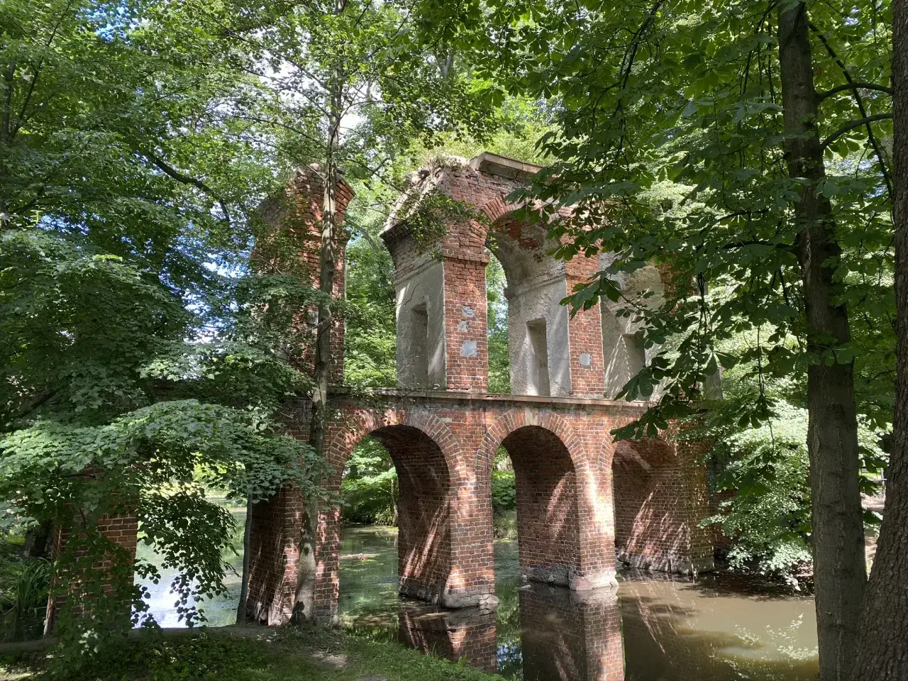 Kamienne łuki i ceglane ruiny tworzą malowniczy park romantyczny w Arkadii, otoczony bujną zielenią i spokojną wodą.