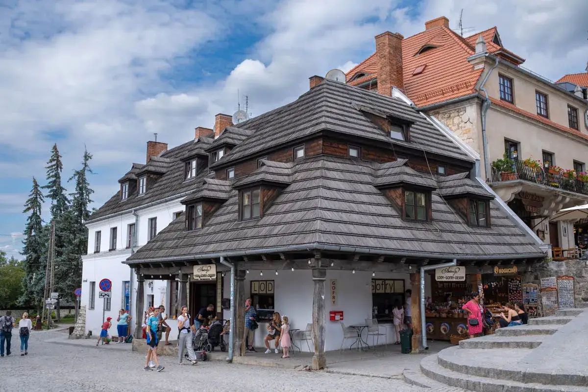 Kazimierz Dolny atrakcje: urokliwy rynek z drewnianymi dachami, lodziarnią i sklepikami. Ludzie spacerują, ciesząc się atmosferą.