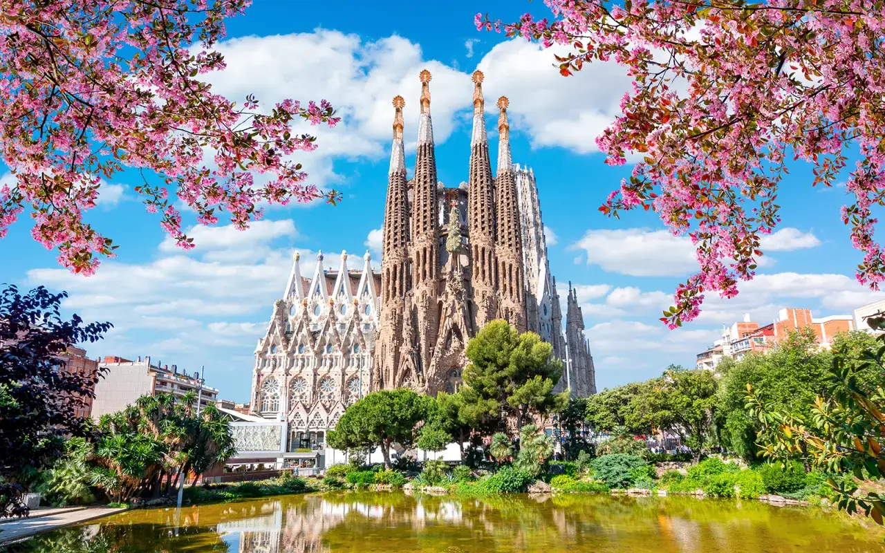Wiosna w Barcelonie. Kwitnące drzewa otaczają majestatyczną Sagrada Familia, odbijającą się w spokojnej tafli wody.