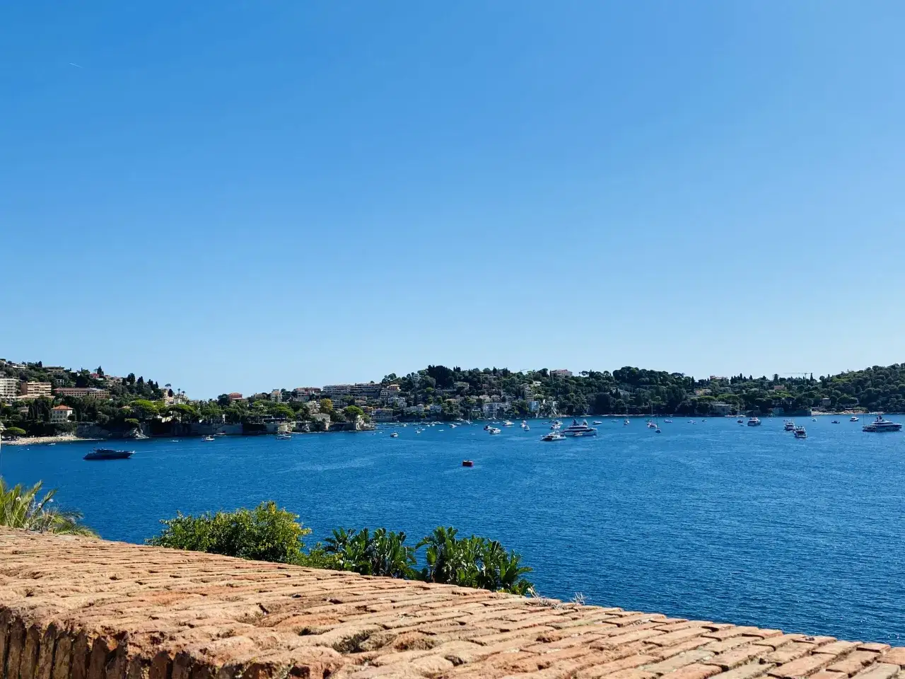 Widok na zatokę w Villefranche-sur-Mer z licznymi jachtami i łodziami na tle zielonych wzgórz i błękitnego nieba.