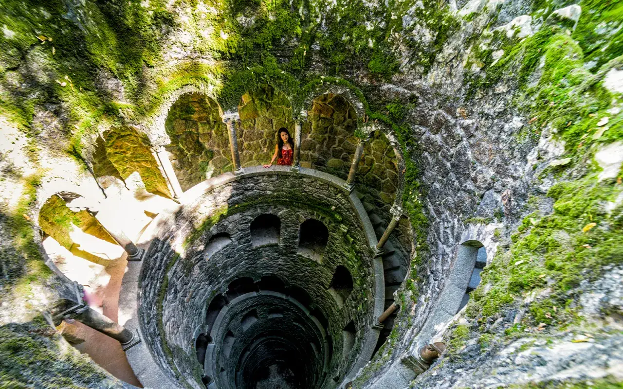 Kobieta na tarasie studni inicjacji w Quinta da Regaleira. Kamienne mury porośnięte mchem, kręte schody w dół.