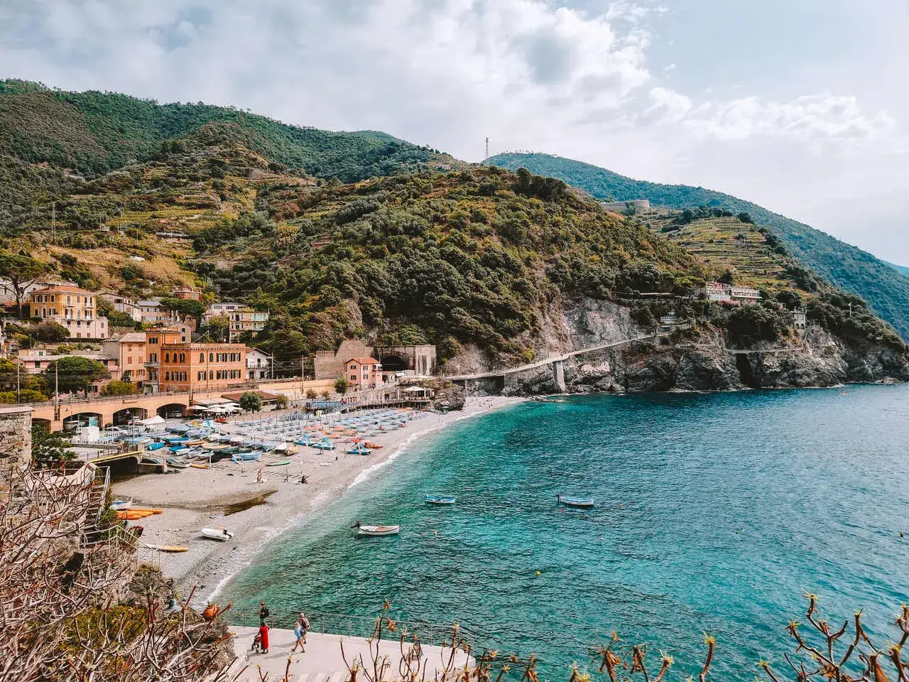 Malowniczy widok na plażę i kolorowe domy w Monterosso al Mare, z zielonymi wzgórzami w tle i turkusowym morzem.