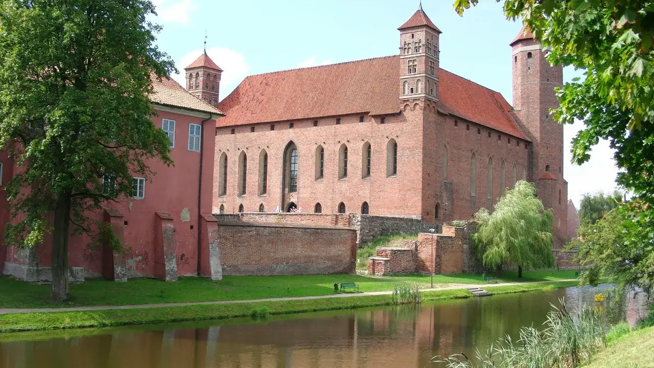 Zamek w Lidzbarku Warmińskim, malownicze atrakcje nad rzeką, z czerwonej cegły, otoczony zielenią.