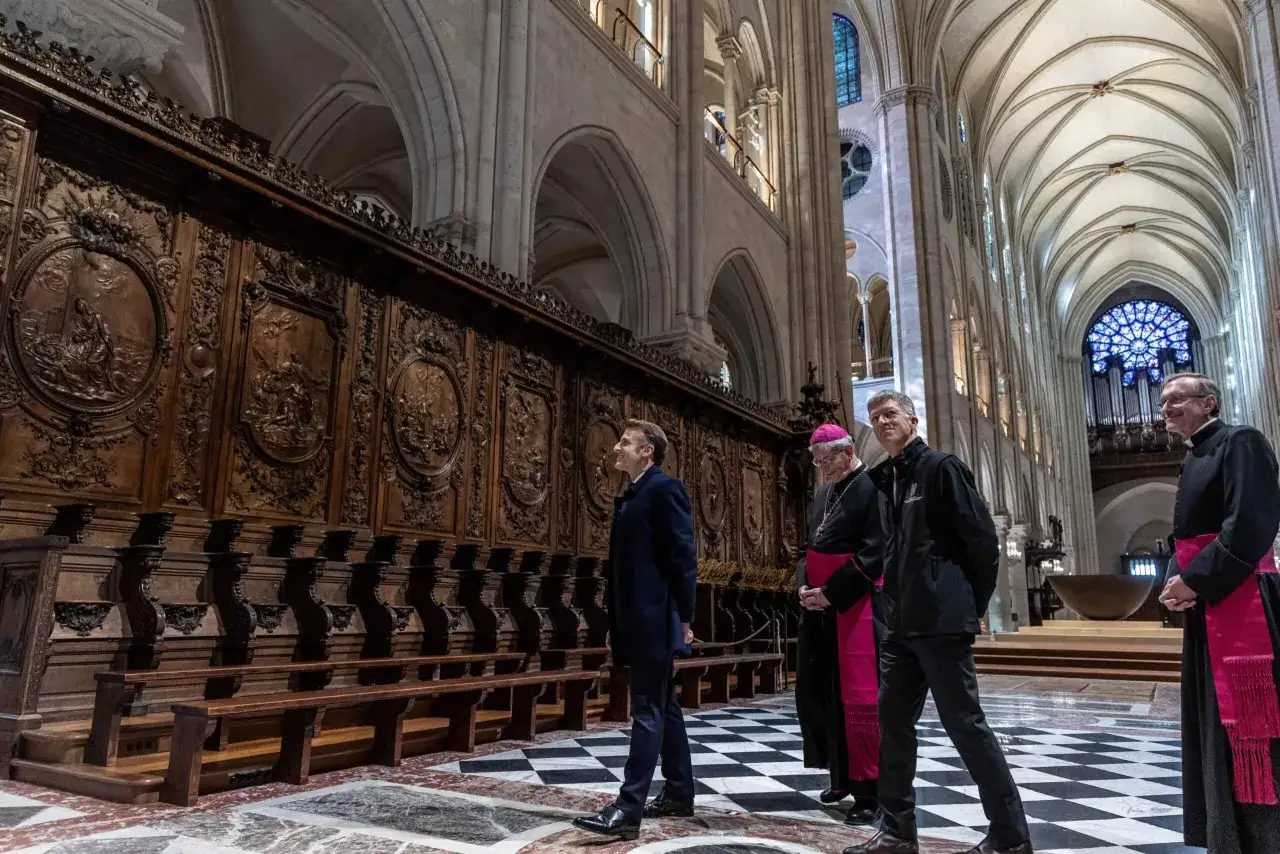 Wnętrze katedry Notre Dame z Macronem i duchownymi podziwiającymi zabytkowe drewniane ławy.