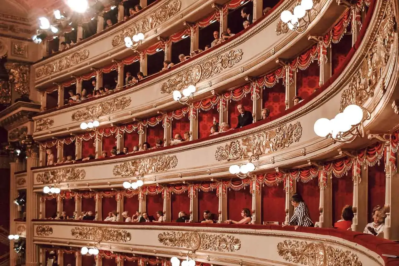 Wnętrze La Scala w Mediolanie, z widownią siedzącą na wielu poziomach, otoczoną złotymi zdobieniami i czerwonymi zasłonami.