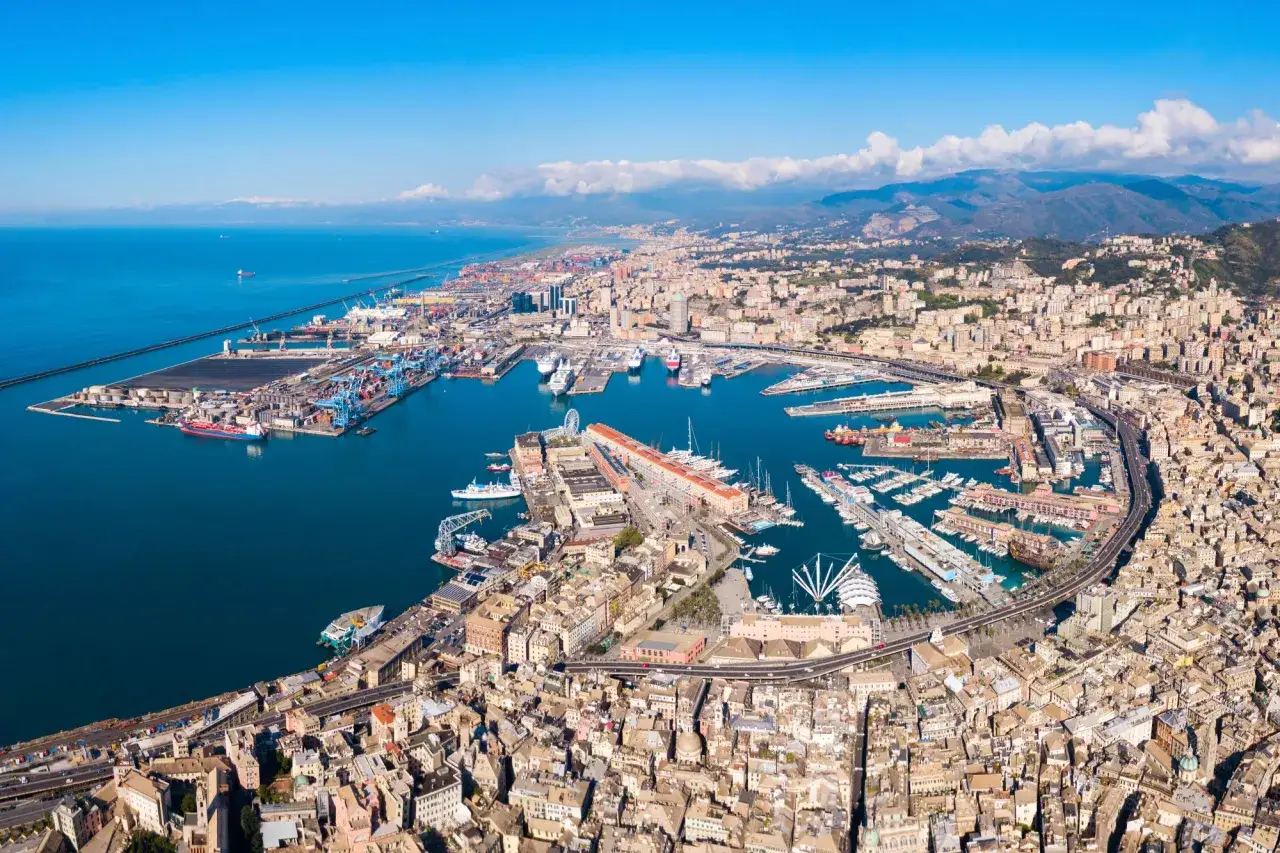 Genua co zobaczyć: rozległy port z dźwigami, statkami i jachtami, otoczony gęsto zabudowanym miastem i górami.