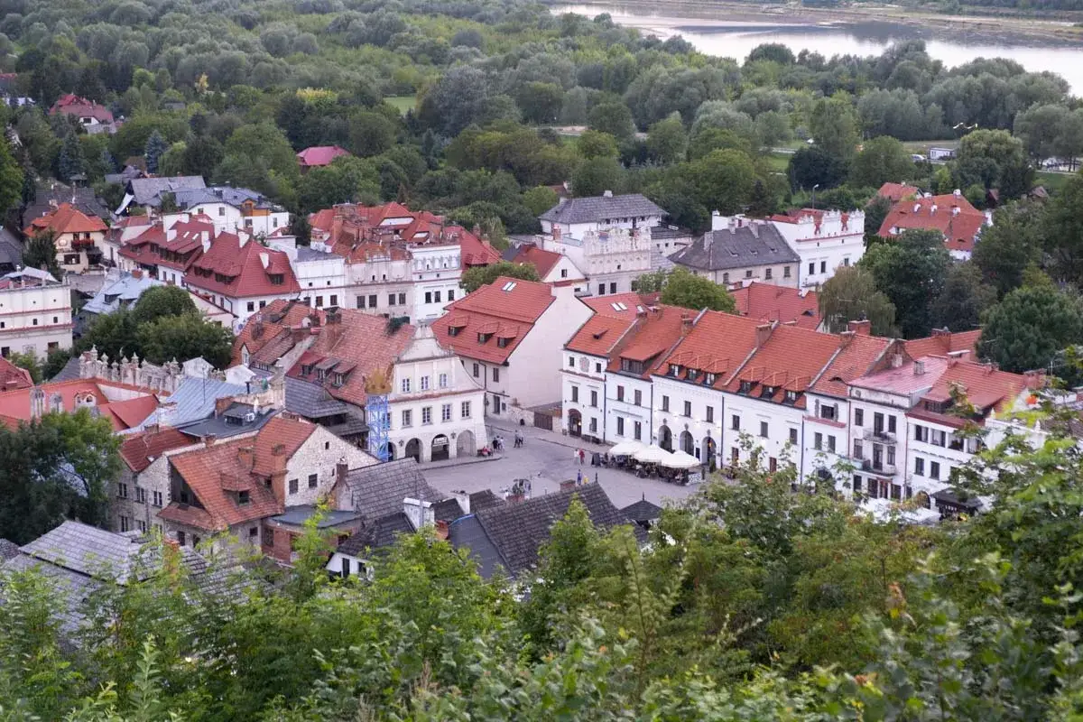 Widok na urokliwe, stare miasto w Kazimierzu Dolnym z charakterystycznymi kamieniczkami i czerwonymi dachami, otoczone zielenią.
