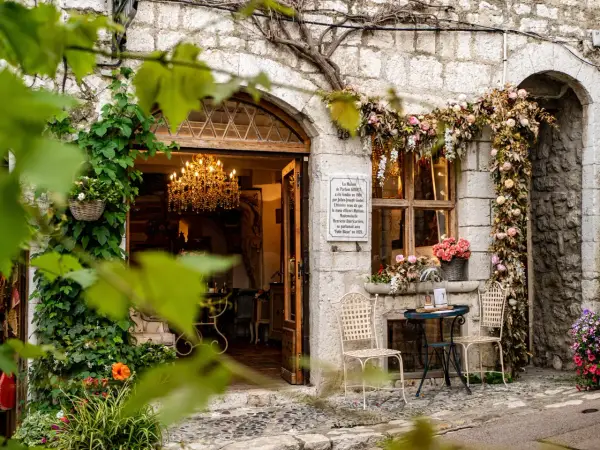 Urokliwa kamienna fasada w Saint-Paul-de-Vence, ozdobiona kwiatami i pnączami. W otwartych drzwiach wisi kryształowy żyrandol.